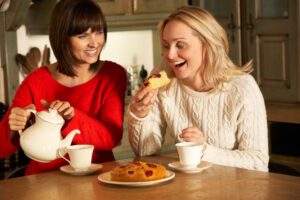 Two white women enjoying tea and cake. One woman has blonde hair and is wearing a white top, the other woman has dark brown hair and is wearing a red top.