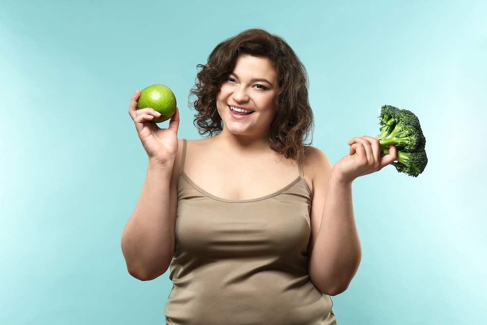 A smiling higher-weight women with brown curly hair, wearing a brown singlet is holding a green apple in her right hand and a head of broccoli in her left hand
