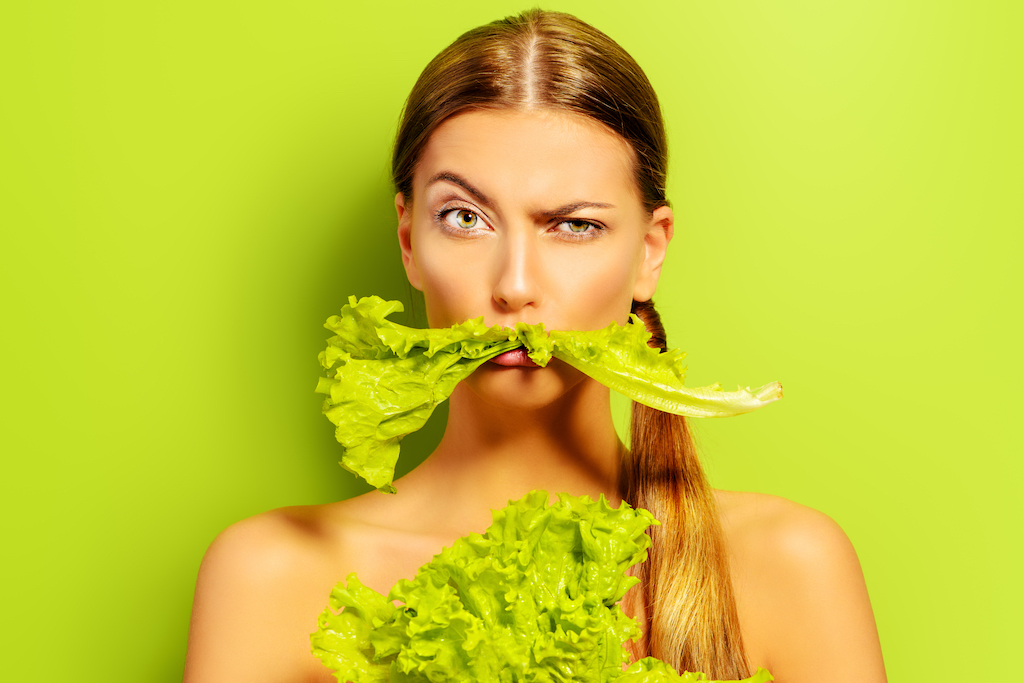 Woman with confused expression on her face. She has a stalk of celery in her mouth and is holding a bunch of celery to her chest