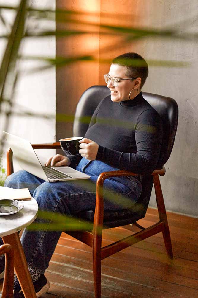 A young person with glasses, with short hair and a nose piercing is sitting in a cafe, drinks coffee and attending supervision on their laptop.