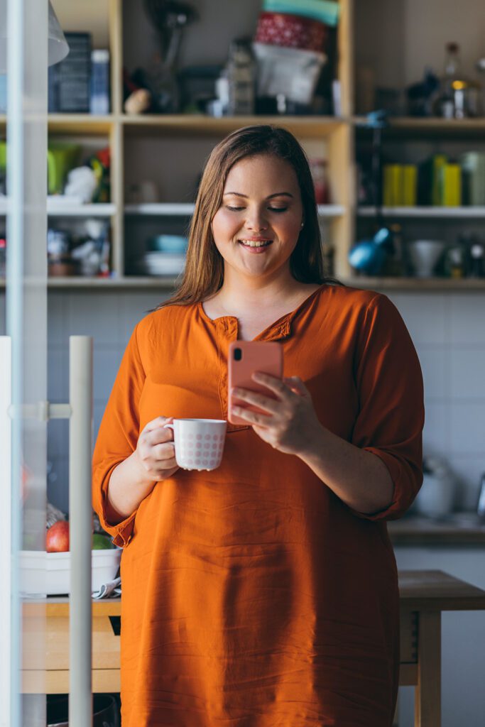 A smiling plus size woman drinking coffee in the morning and making an appointment for weight inclusive nutrition counselling on her smartphone in the kitchen